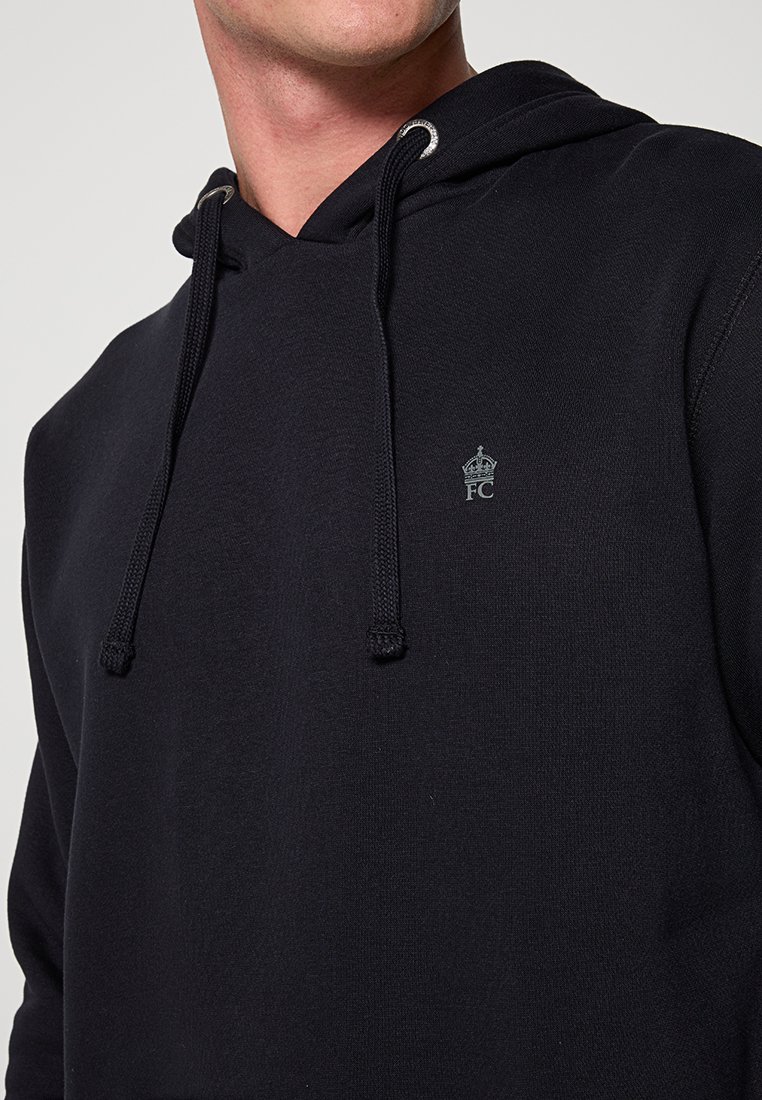 Ein Mann trägt einen schwarzen Hoodie mit Kordeln und einem kleinen Kronenlogo mit „FC“ auf der Brust vor einem schlichten Hintergrund.
