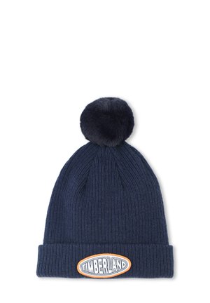 Bonnet en tricot bleu marine avec un design à côtes texturées et un grand pompon en fausse fourrure sur le dessus. Il présente un patch logo Timberland bien en vue à l'avant.