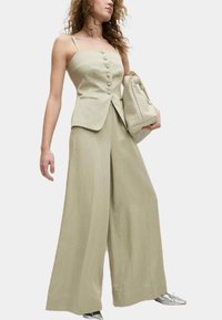 Hellgrünes, ärmelloses Peplum-Top mit Knöpfen und zwei Taschen, kombiniert mit weit geschnittenen Hosen. Das Model trägt eine strukturierte Handtasche.