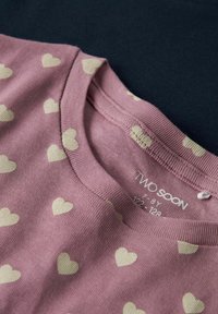 Nærbillede af lyserød børnetrøje med cremefarvet hjerteprint og rund halsudskæring, størrelsesmærke viser "TWO SOON 7-8Y 122-128".