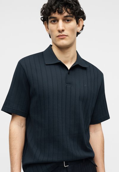 Νέος άνδρας που φοράει μαύρο κοντομάνικο ριπ πικέ πόλο με γιακά, στέκεται μπροστά σε απλό φωτεινό φόντο.