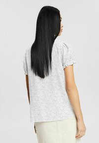Mujer con cabello largo y negro que lleva una blusa blanca de manga corta con lunares negros y una falda de color claro, de espaldas.