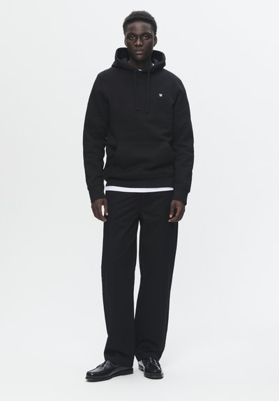 Sweatshirt à capuche noire avec poche avant et logo brodé, associé à un pantalon noir ample et des chaussures noires. Design simple et monochrome.