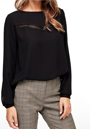 Schwarze Bluse mit langen Ärmeln und Spitzendetails am Ausschnitt, kombiniert mit karierten Hosen in Beige- und Brauntönen. Glatte Stofftextur.