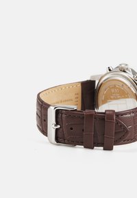 Correa de reloj de cuero marrón con un patrón de cocodrilo en relieve, hebilla de metal plateado y una caja de metal redonda visible en la parte posterior.