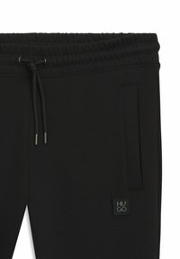 Schwarze Jogginghose mit elastischem Bund und Kordelzug, Seitentasche und gummiertem Logo-Patch auf der Vorderseite. Glatte Stofftextur.