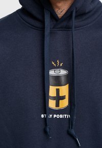 Sudadera de color azul marino oscuro hecha de algodón, con un gráfico de batería en negro y amarillo con rayos sobre ella y el texto "MANTENTE POSITIVO."