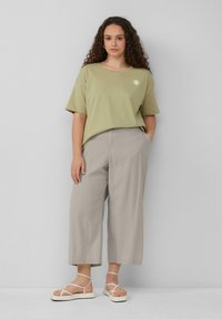 Olivenfarbene Baumwoll-T-Shirt mit kurzen Ärmeln und Sonnen-Grafik, kombiniert mit hellgrauen weit geschnittenen Hosen und beigen Riemchensandalen.