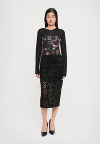 Woman wearing a black long-sleeve top with a floral chest panel, a black lace midi skirt, and black pointed-toe heels, standing against white.