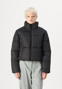 Černá vycpaná bunda s vysokým límcem, zapínáním na zip a vodorovným prošíváním. Nositá přes světle šedé kalhoty. Hladká textura.