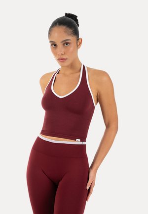 Kobieta o ciemnych włosach związanych w kok, ubrana w bezrękawnikowy bordowy crop top i dopasowane legginsy z wysokim stanem z białym wykończeniem, stojąca na białym tle.