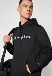 Felpa nera con cappuccio e coulisse, con un audace logo "Champion" bianco e colorato. Include una tasca frontale e un accento del logo sulla manica.