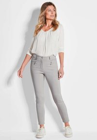 Lichtgrijze slim-fit broek met zilveren ritszakken, gecombineerd met een witte blouse met een ronde halslijn en driekwart mouwen.