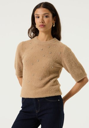 Frau trägt einen beigefarbenen, flauschigen Kurzarm-Pullover mit kleinen silbernen Nieten, dunkle Jeans und goldene Creolen und steht vor weißem Hintergrund.