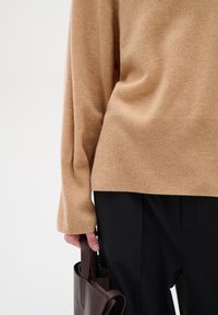 Pulover bej din tricot cu tiv și mansete ribbate, asortat cu pantaloni negri. O geantă din piele maro este ținută în mână.