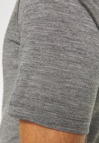 Κοντινό πλάνο στο χέρι ενός ατόμου που φοράει γκρι πλεκτό πουκάμισο με κοντό μανίκι, δείχνοντας την  υφή του υφάσματος και τις λεπτομέρειες της ραφής.