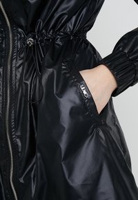 Černá nepromokavá bunda s zipovým uzávěrem, elastickými manžetami a staženým pasem se boční kapsou. Hladký, lesklý povrch.