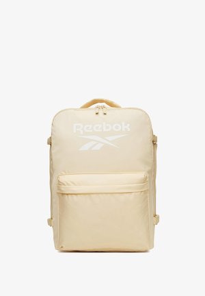 Mochila beige de Reebok con asa superior, bolsillo frontal con cremallera y logo blanco en el panel frontal sobre fondo blanco.