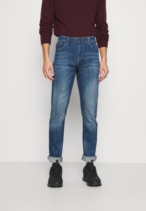 Jeans de mezclilla azul oscuro con un fit ajustado y cónico, puños enrollados y costuras en contraste. Combinados con zapatillas negras.