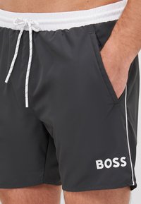 Bañador negro con un cinturón elástico blanco, cordón ajustable, bolsillos laterales y un logo blanco en la parte inferior derecha. Textura suave.