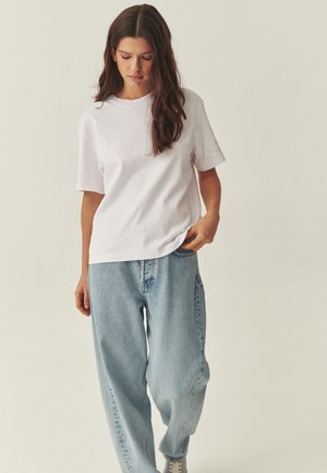 Woman with long brown hair wearing a plain white t-shirt and loose light blue jeans standing against a neutral background.