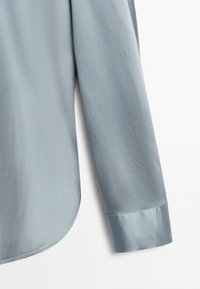 Manica di camicia a manica lunga azzurra chiaro con polsino largo e orlo curvo su sfondo bianco.
