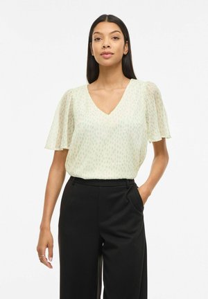 Kvinde med langt mørkt hår iført en lys, prikket, kortærmet bluse og sort bukser med høj talje, den ene hånd i lommen, foran hvid baggrund.