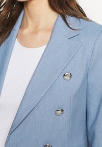 Ljusblå skräddarsydd kavaj i strukturerad tyg, med dubbelknäppt design och reflekterande silverknappar.