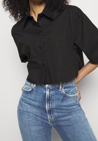 Camisa negra de manga corta con botones y bolsillo, que presenta un logotipo redondo en el pecho. Combinada con jeans de tiro alto de color azul claro.