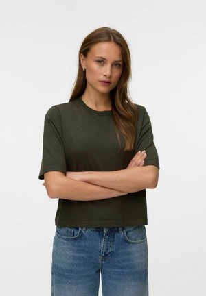 Donna con capelli castani lunghi indossa una maglietta verde scuro a maniche corte e jeans blu, in piedi con le braccia incrociate davanti a uno sfondo semplice.