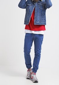 Jeansjacka, röd topp, vit lagertröja, blå jeans, grå sneakers med röda accenter. Avslappnad outfit med distinkta färglager och texturer.