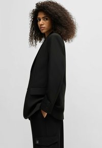 Blazer nero su misura con spalle strutturate, design monopetto e dettagli delle tasche. Abbinato a pantaloni neri coordinati.
