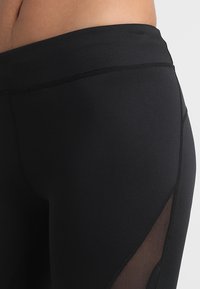 Lähikuva henkilön vyötäröstä ja lantion alueesta, joka on pukeutunut mustiin legginsseihin, joissa on verkkopaneelit yksityiskohtien osalta sivuilla.