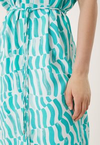 Robe turquoise sur fond blanc, avec des motifs audacieux de coups de pinceau et une ceinture nouée à la taille. Texture du tissu douce visible.