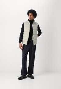 Light beige puffer vest, ribbed texture, zippered front, worn over a black turtleneck. Black pinstriped wide-leg pants and black shoes.