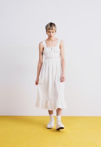Robe midi blanche avec décolleté carré, corsage froncé et ourlet à volants. Portée avec des baskets blanches sur un sol jaune.