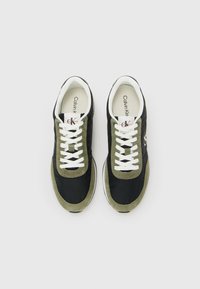 Een paar zwart en olijfgroene Calvin Klein sneakers met witte veters, van bovenaf gezien op een witte achtergrond.
