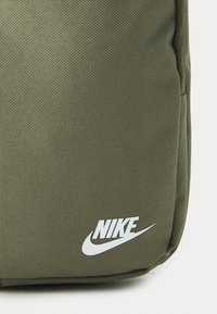 Olīvu zaļš mugursoma ar teksturētu audumu, ar izteiktu baltu Nike logotipu un izliekta Nike swoosh akcentu apakšā.