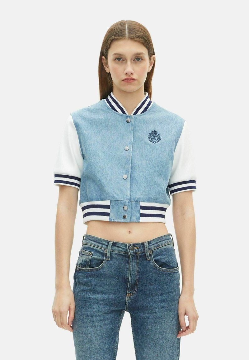 Skórzana kurtka jeansowa z białymi rękawami, granatowo-białym, prążkowanym kołnierzem, haftowanym logo i zatrzaskami. W komplecie z niebieskimi dżinsami.