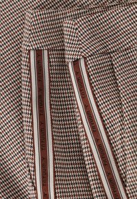 Textilie s hnědo-krémovým vzorem houndstooth s texturovaným povrchem, doplněná kontrastní pruhovanou stuhou podél okrajů.