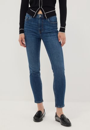 Persona che indossa jeans skinny blu scuro, mocassini neri e un top nero a maniche lunghe con piping bianco, in piedi davanti a uno sfondo semplice.
