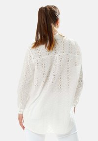 Long-sleeve white shirt made of textured fabric, featuring cut-out patterns, a back yoke, and buttoned cuffs. The fit is relaxed.