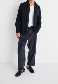 Svart skjorta med knappar och två bröstfickor, i kombination med lös mörkgråa jeans och svarta loafers, mot en ljus bakgrund.