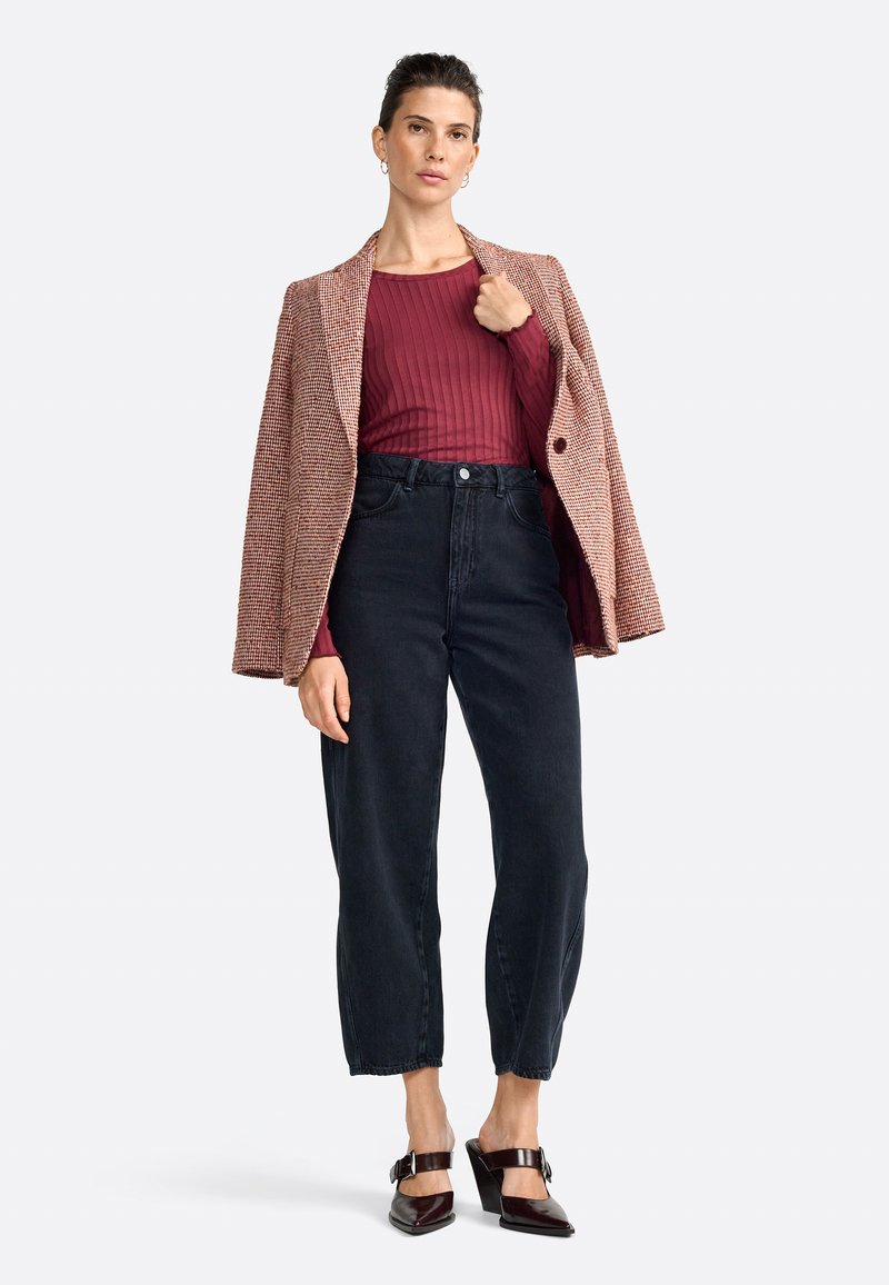 Haut côtelé rouge sous un blazer marron à motif pied-de-poule, associé à un jean foncé taille haute et des chaussures à talons brillantes bordeaux.