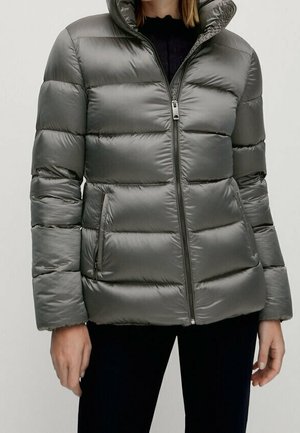 Femme portant une doudoune matelassée gris brillant avec fermeture éclair à l'avant et poches latérales zippées, associée à un pantalon noir.