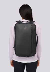 Person med långt mörkt hår som bär en ljuslila tröja och en svart ryggsäck med en vertikal dragkedjeficka och ett cirkulärt märke.