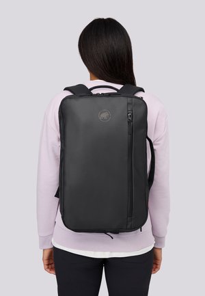 Persona con lunghi capelli scuri che indossa un pullover viola chiaro e uno zaino nero con una tasca verticale con zip e un logo circolare.
