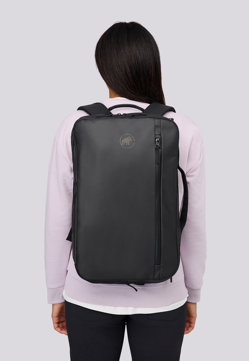 Person med långt mörkt hår som bär en ljuslila tröja och en svart ryggsäck med en vertikal dragkedjeficka och ett cirkulärt märke.