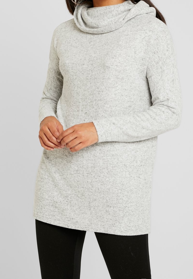 Femme portant un pull à col bénitier manches longues de couleur gris clair, les mains jointes, assorti à un pantalon noir sur un fond uni.