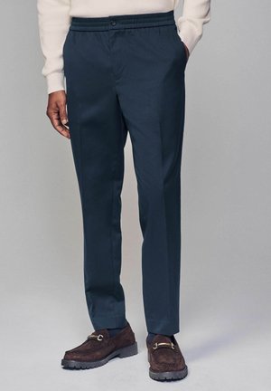 Navy blue trousers with a relaxed fit, elastic waistband, button closure, and side pockets. Worn with brown suede shoes.